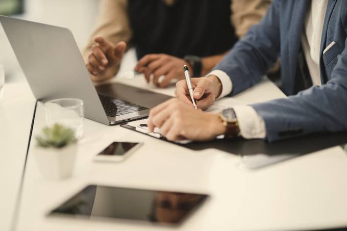 Two people at a desk, one writing on a pad with a pen while the other's hands rest open on the table.