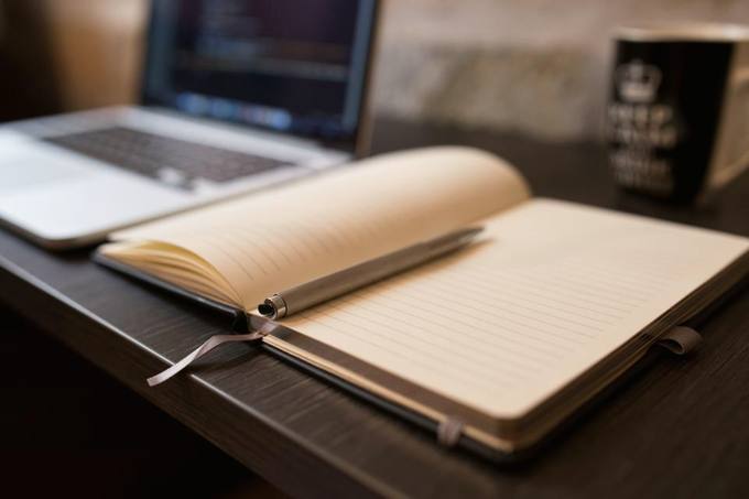 An open notebook with a pen in the fold on a desk, a laptop and a dark mug behind it, lit by warm lamplight.