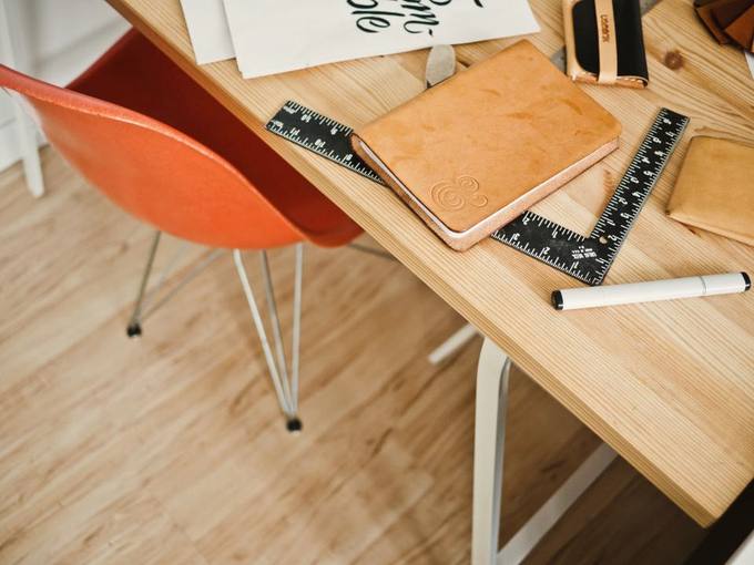 A pale wooden desk seen from above with a tan leather notebook, a steel ruler and a marker.