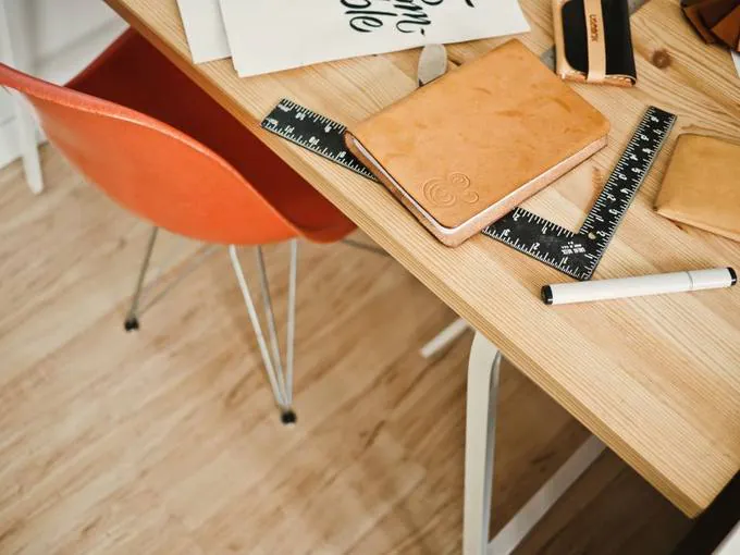 A pale wooden desk seen from above with a tan leather notebook, a steel ruler and a marker.